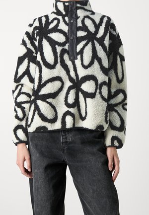 Blød fleece-trøje med sort abstrakt blomstermønster på hvid baggrund, knappet høj krave, båret med mørke vaskede jeans.