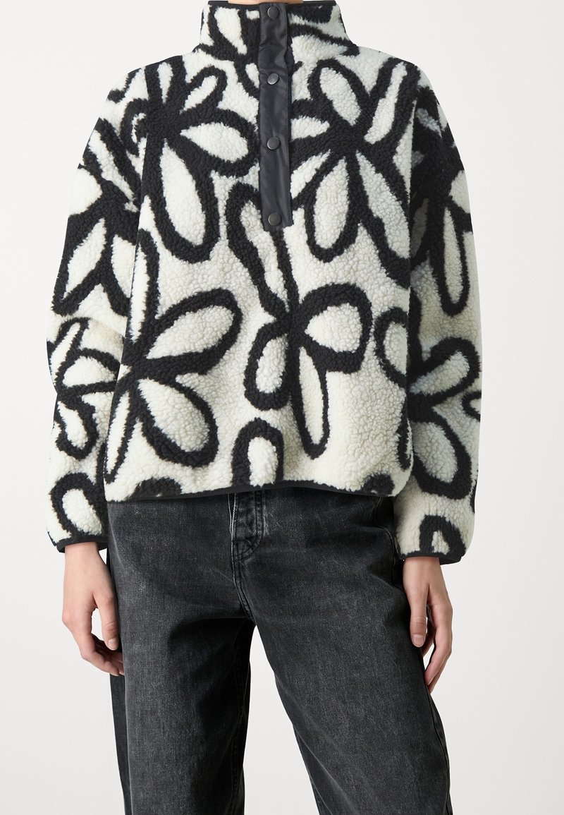 Pull en polaire douce avec motif floral abstrait noir sur fond blanc, col montant boutonné, porté avec un jean délavé foncé.