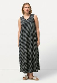 Maxi šaty černé barvy s bílým listovým vzorem, bez rukávů, s výstřihem do V, volný střih a splývavá látka. K tomu jsou nošeny béžové šlapky.