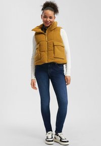 Frau steht da und trägt eine senfgelbe Cord-Pufferweste, ein weißes Langarmshirt, dunkelblaue Skinny-Jeans und weiße Sneakers mit schwarzen Akzenten.