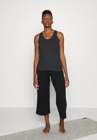 Top de algodão preto sem mangas com decote redondo, combinado com culotes pretos de corte solto. Modelo em pé sobre uma superfície cinza texturizada.