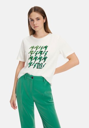 Weißes T-Shirt mit kurzen Ärmeln und grünem Grafikdruck, der abstrakte Formen zeigt. Kombiniert mit grünen, maßgeschneiderten Hosen mit einem Knopfdetail vorne.