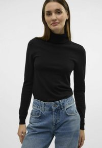 Črna turtleneck pulover s dolgimi rokavi, izdelan iz gladkega materiala, v kombinaciji z svetlo modrimi visokim pasom kavbojkami, ki imajo sprednje žepke.