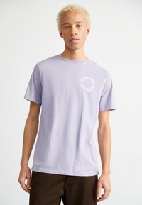 Camiseta de algodón de color púrpura claro con cuello redondo y mangas cortas. Presenta un gráfico circular blanco en el lado izquierdo del pecho. Textura suave.