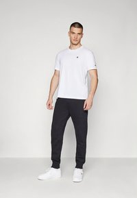 Fehér pamut póló kis logóval a mellkason, fekete melegítőnadrággal és fehér tornacipővel, semleges háttér előtt.