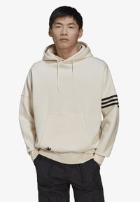 Hoodie color crema con tasca frontale, accenti neri a tre strisce sul maniche sinistro e un piccolo logo nero vicino all'orlo.