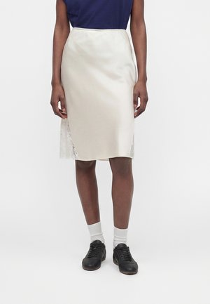 Žena nosi krémovú saténovú sukňu po kolená s čipkovými stranami, námornícky modré tričko, biele ponožky a čierne šnurovacie topánky.