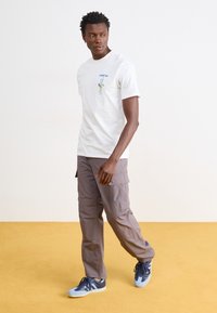 T-shirt bianco a maniche corte con un grafico in stile cartone animato, abbinato a pantaloni cargo grigi e scarpe da ginnastica blu. Il pavimento è in moquette gialla.