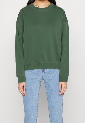 Person iført en ensfarvet mørkegrøn langærmet sweatshirt og lyseblå jeans, stående mod en ensartet hvid baggrund.