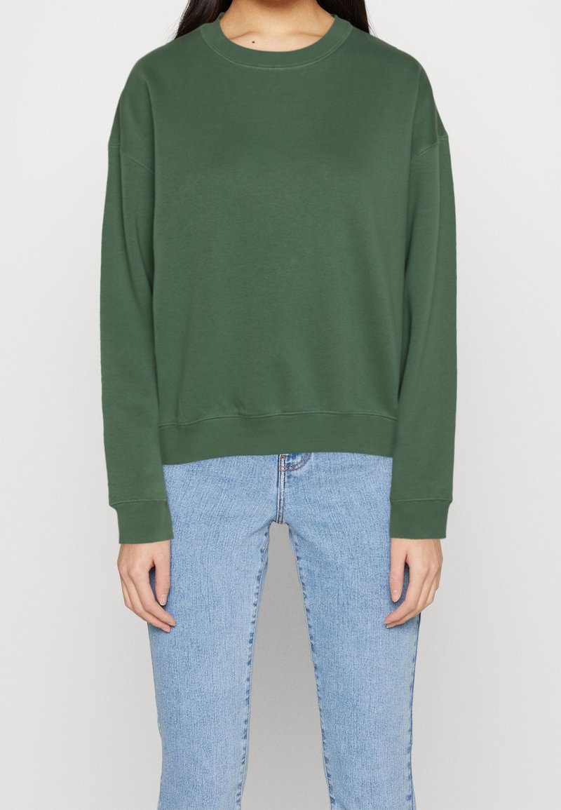 Personne portant un sweat-shirt uni vert foncé à manches longues et un jean bleu clair, debout devant un fond blanc uni.