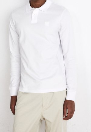 Biała koszulka polo z długim rękawem, kołnierzykiem zapinanym na guziki oraz małym kwadratowym logo. Materiał wydaje się gładki i lekko teksturowany.