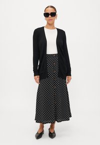 Cardigan preto sobre uma blusa branca, combinado com uma saia preta com bolinhas brancas e botões. Os acessórios incluem óculos de sol pretos e sapatos rasos.