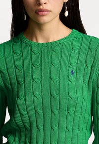 Kvinna i en grön kabelstickad tröja med en liten blå broderad logotyp på bröstet och vita snäckor som hänger som örhängen.