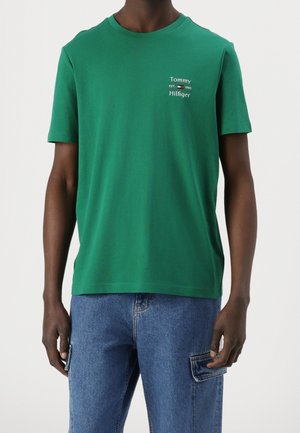 Mężczyzna ubrany w zieloną koszulkę Tommy Hilfiger z logo na piersi oraz niebieskie dżinsowe spodnie cargo na białym tle.