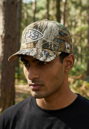 Mann mit Camouflage-Kappe der Kansas City Chiefs und schwarzem Hemd steht in einem Wald mit verschwommenen Bäumen im Hintergrund.