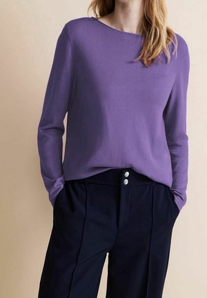 Femme portant un pull violet à manches longues rentré dans un pantalon noir taille haute avec deux boutons visibles, les mains dans les poches.