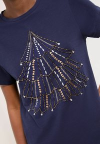 Tmavě modré bavlněné tričko s dekorativním vzorem stromu vyrobeným ze zlatých a stříbrných korálků, s rámovanými vlnitými okraji a různorodými texturami.