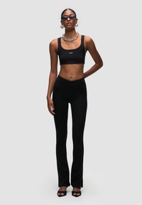 Zwart geribbeld crop top gecombineerd met hoge zwarte wijde broek. De outfit heeft strakke lijnen en een gladde textuur, benadrukt met kettingen accessoires.