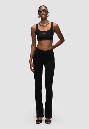 Top corto a costine nero abbinato a pantaloni neri a vita alta e svasati. L'outfit presenta linee eleganti e una texture liscia, accentuato da accessori a catena.