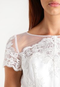 Bílá šaty s krajkovým lemem na průsvitných síťovaných rukávech. Hladká tkanina s květinovými vzory, kulatý výstřih a knoflíková detaile na přední straně.