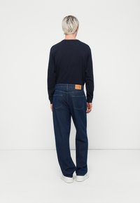 Dark blue jeans with a relaxed fit, featuring two back pockets, a leather brand patch, and clean stitching. Paired with a long-sleeve black top.