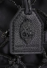 Il tag in pelle nera testurizzata presenta un design in rilievo di una mano hamsa decorata con cristalli neri. Ha un bordo liscio e un anello per il fissaggio.