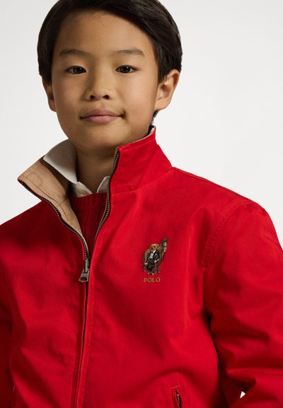 Piros cipzáros dzseki bézs gallérral, hímzett logóval a mellrészen és texturált anyagból készült kivitelben.