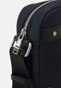 Borsa in nylon blu navy con una cerniera argentata, dettagli in pelle nera e un gancio metallico con strisce rosse, bianche e blu. Superficie liscia e forma strutturata.