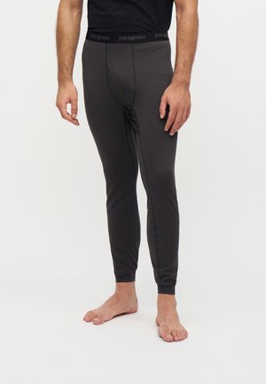 Homme portant des leggings longs de sous-vêtements noirs Patagonia et un t-shirt noir, debout pieds nus sur un fond blanc.