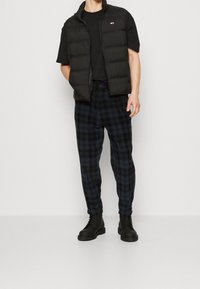 Gilet imbottito nero sopra una maglietta nera, abbinato a pantaloni a quadri blu navy e stivali neri con lacci. L'insieme presenta un design semplice e informale.