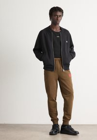 Young man standing with hands in pockets wearing black zip-up jacket, black T-shirt, brown jogger pants, and black sneakers on plain background.