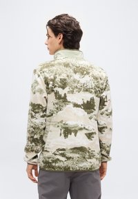 Άνδρας που φοράει φλις μπουφάν με καμουφλαρισμένο μοτίβο σε πράσινο και μπεζ, με ψηλό γιακά, που φαίνετ αι από πίσω μπροστά σε απλό φόντο.