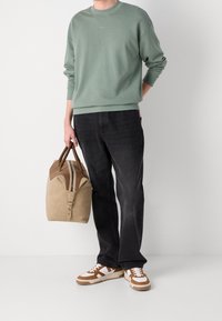 Felpa verde, jeans neri, scarpe da ginnastica marroni con dettagli bianchi. Tiene un borsone beige con dettagli in pelle marrone e cerniera.