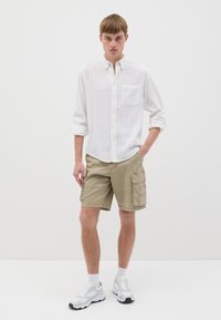 Camicia bianca a maniche lunghe con bottoni e taschino sul petto, abbinata a pantaloni cargo beige con tasche laterali, indossati con sneakers bianche.