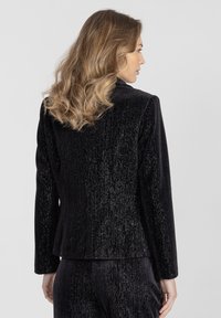 Czarny aksamitny blezer z pionową fakturą, z konstrukcyjnymi ramionami i długimi rękawami. Materiał ma delikatny połysk, który podkreśla jego wizualny urok.