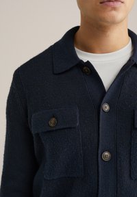 Marinblå stickad skjortjacka med en texturerad yta, utrustad med krage, knäppning med knappar och två bröstfickor med knäppbara flikar.