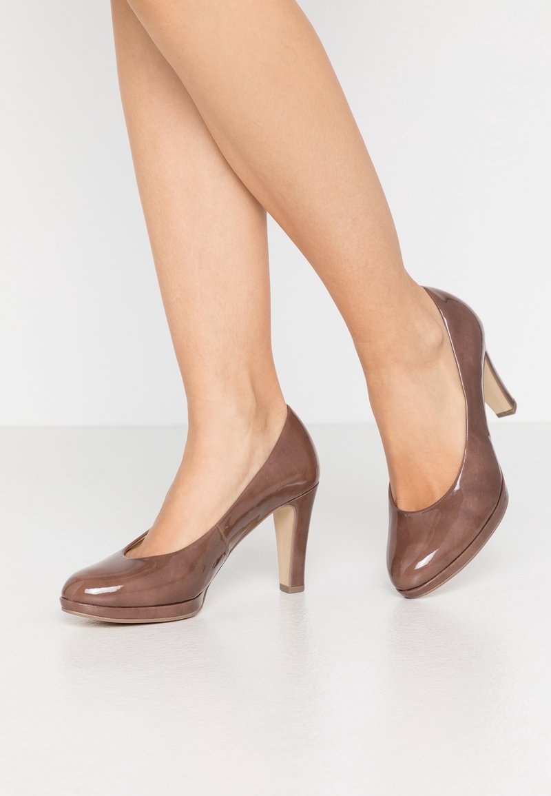 Femme portant des chaussures à talons fermés brillantes marron, se tenant sur un sol blanc lisse avec un pied légèrement levé.