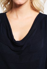 Marinblå topp med draperad halsringning, texturerad tyg med växlande horisontella ränder och en slät finish.