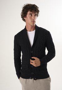 Zwarte gebreide cardigan met een kabelpatroon, voorzien van een sjaalkraag, vijf knopen en een voorzak. Gedragen over een witte blouse.