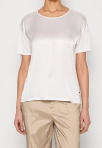 Biała koszulka z krótkim rękawem o okrągłym dekolcie, miękka w dotyku i delikatnie połyskująca, w zestawieniu z jasnobeżowymi, delikatnie połyskującymi, luźnymi spodniami.