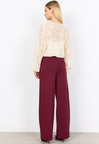 Top in pizzo color crema con maniche lunghe e vaporose, abbinato a pantaloni a vita alta, larghi e di un intenso color bordeaux. Texture liscia e vestibilità ampia.