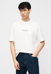 Νεαρός άνδρας με λευκό μπλουζάκι Hollister και σκούρο μπλε τζιν, στέκεται με το ένα χέρι στην τσέπη μπροστά σε απλό ανοιχτό φόντο.