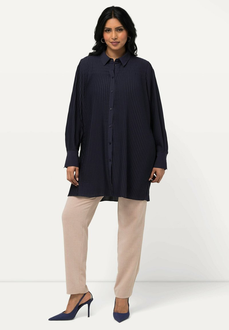 Camicia tunica a pieghe di colore navy con maniche lunghe e chiusura a bottoni, abbinata a pantaloni attillati beige e décolleté blu a punta.
