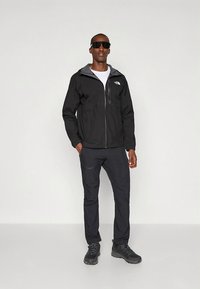 Schwarze wasserdichte Jacke mit grauen Akzenten, Reißverschlusstasche auf der Brust; kombiniert mit schwarzer Hose und robusten schwarzen Wanderschuhen.