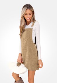 Bauchedress in Tan mit Schulterträgern, Taschen und Knopfdetails. Kombiniert mit einer weißen Handtasche und braunen Stiefeln.