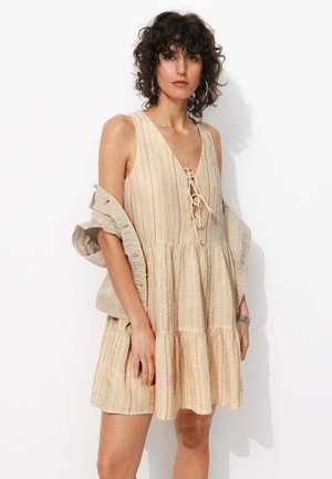 Robe sans manches à rayures en beige clair avec des accents orange, présentant un décolleté en V et un détail à nouer. Jupe fluide et superposée avec une coupe décontractée.