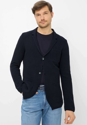 Man die een getextureerd marineblauw colbert draagt, een zwart overhemd en lichtblauwe jeans, staand tegen een witte achtergrond met één hand in de zak.