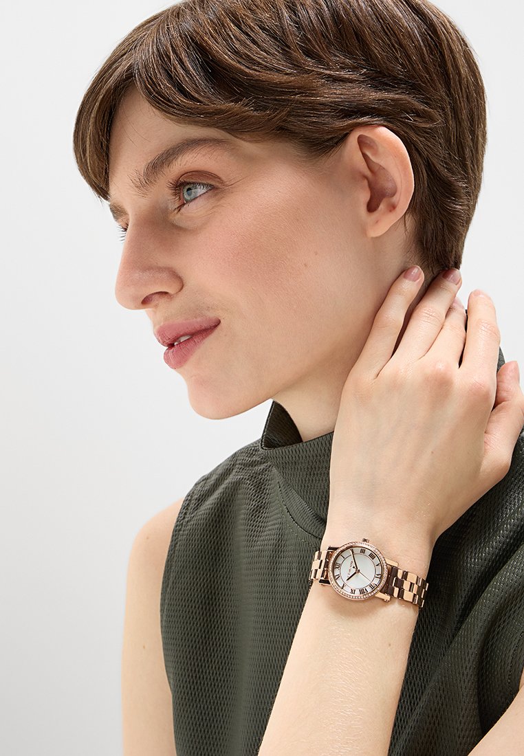 Femme aux cheveux bruns courts portant un haut sans manches vert foncé, affichant une montre-bracelet en or rose avec un cadran blanc et des chiffres romains.