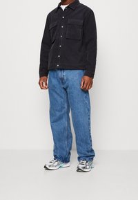 Černá kordurová bunda se cvoky a dvěma náprsními kapsami, v kombinaci s světlými modrými volnými džíny a stříbrnými sportovními botami s tyrkysovými akcenty.