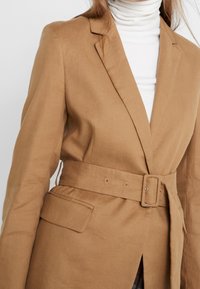 Brązowy lniany żakiet z paskiem w talii, klapami i bocznymi kieszeniami. Teksturowany materiał z dopasowanym krojem i detalami z zapięciem na guziki.